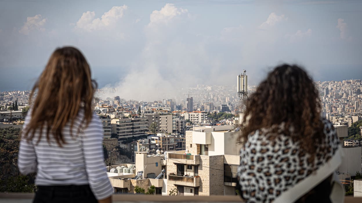 Two women look out over a city skyline filled with buildings and a plume of smoke.