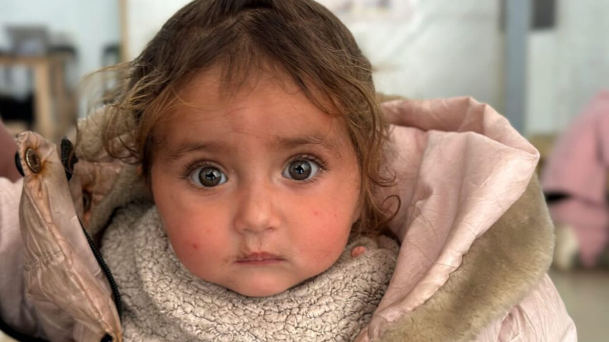 Close up of a young child with big eyes wearing a pink jacket and gray sweater.