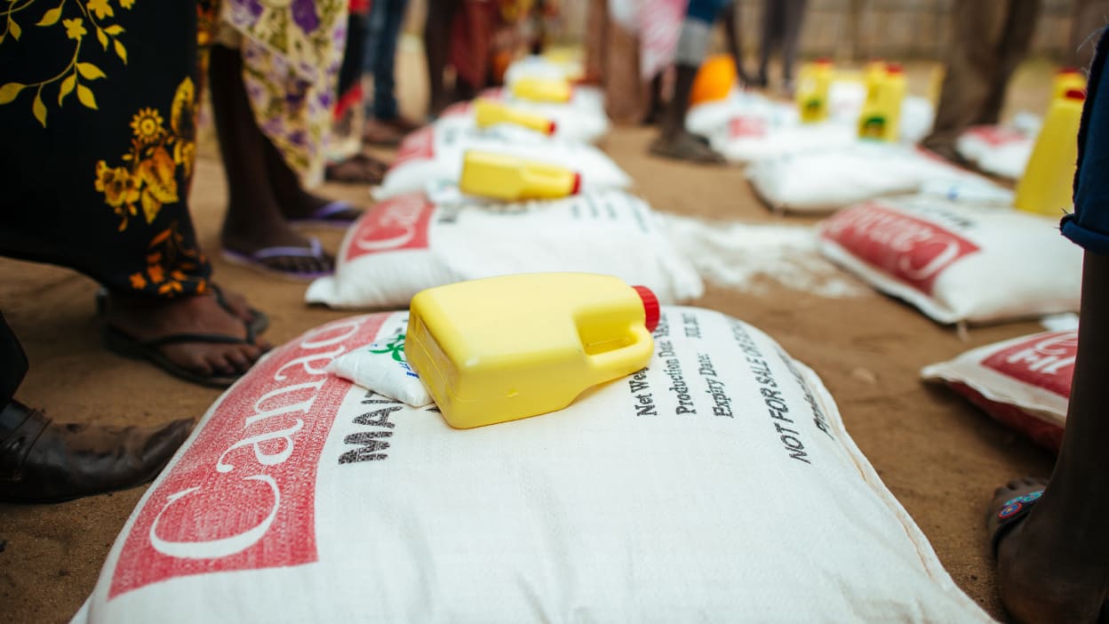 Several people stand around a delivery of food supplies, including a white bag of maize flour and a yellow bottle of oil.