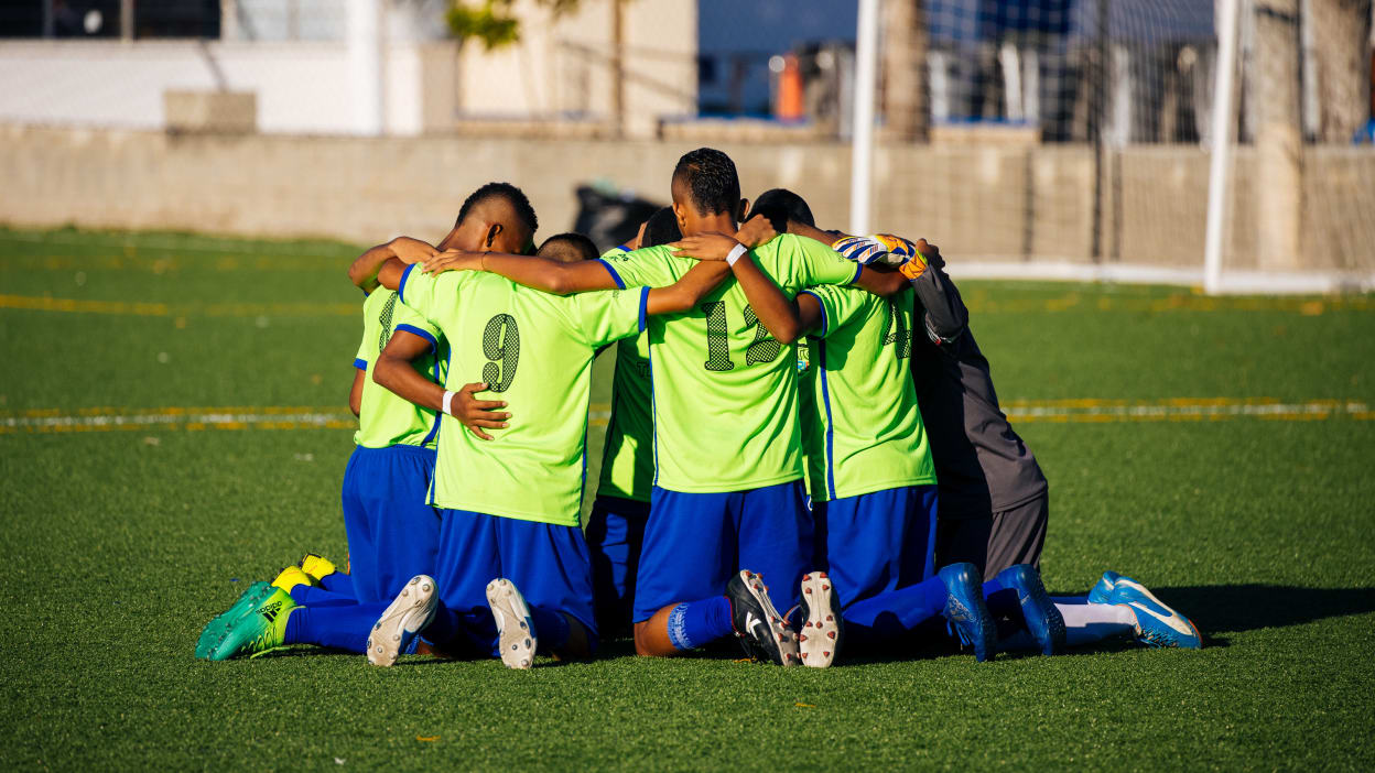 Soccer players in bright green jerseys huddle together on their knees on a green turf field.