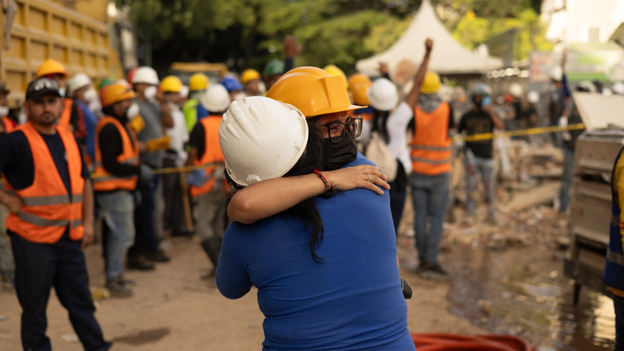 Two people in hard hats hug amidst a crowd of rescue workers at a debris-filled earthquake disaster site.