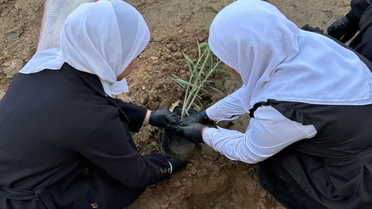 Two girls wearing headscarves and black gloves carefully plant a small green plant into the soil.