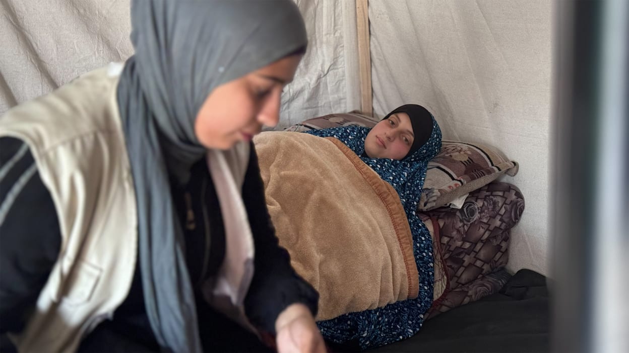 Woman in grey hijab tends to a sick woman resting under a blanket in a humanitarian tent.