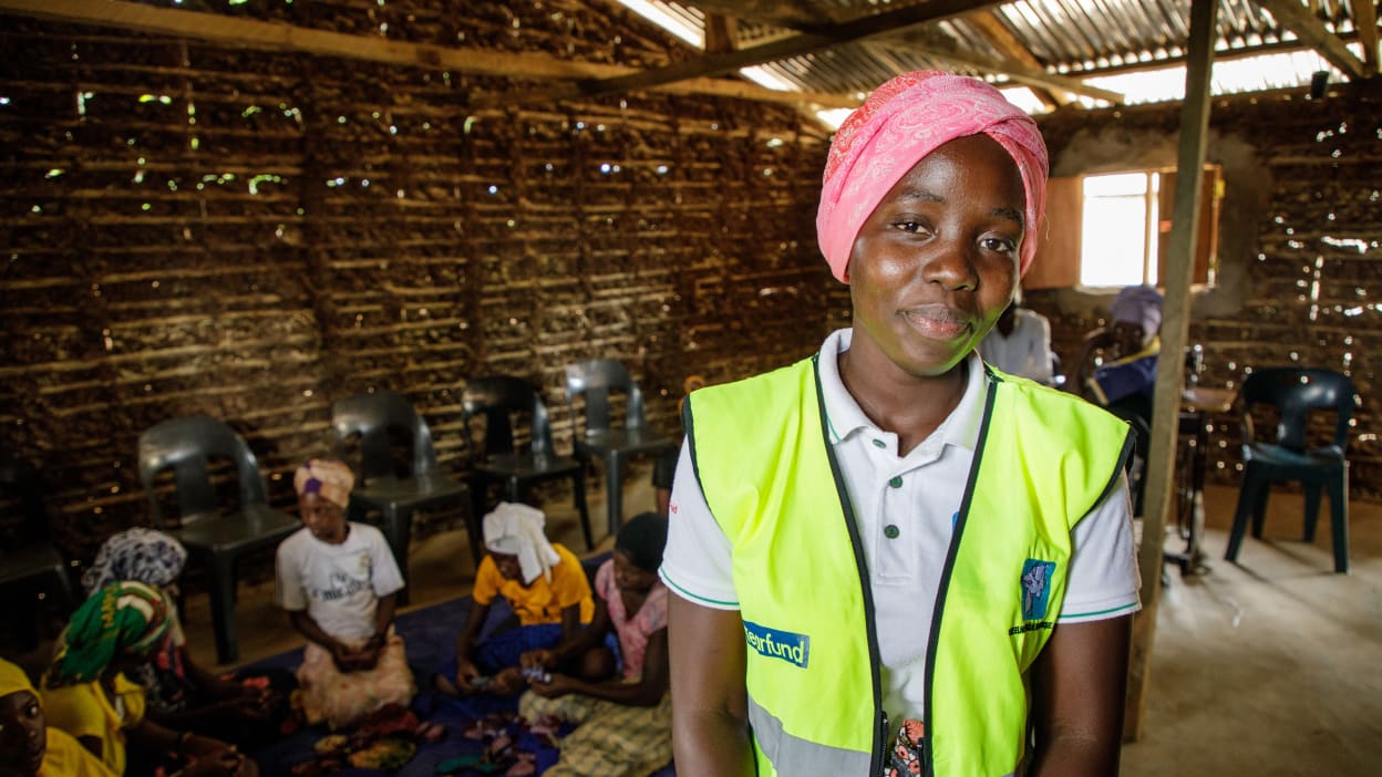 A young Black woman wearing a pink headscarf and a high-visibility yellow vest smiles at the camera. She stands indoors in a room with woven walls and a corrugated metal roof, with other women seated on the floor and on chairs behind her.