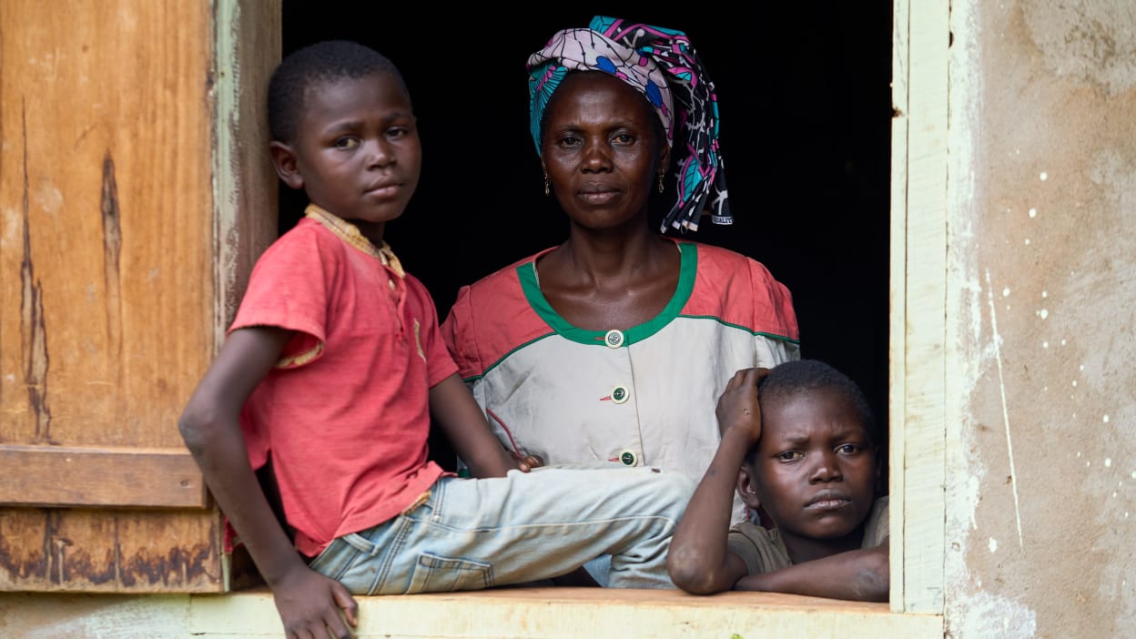 Woman and two children look out of a wooden window with dark background, somber expression.