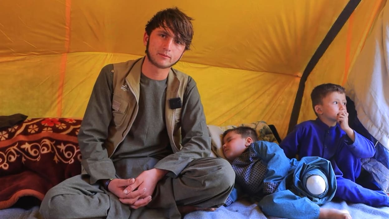 Man sits with two children inside a yellow tent. One child sleeps beside him.