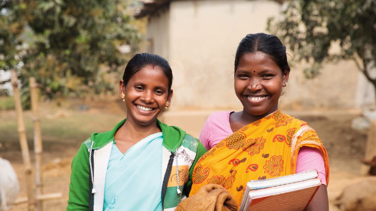 Duas mulheres de um grupo comunitário de Bangladesh sorrindo para a câmera, uma delas segurando livros