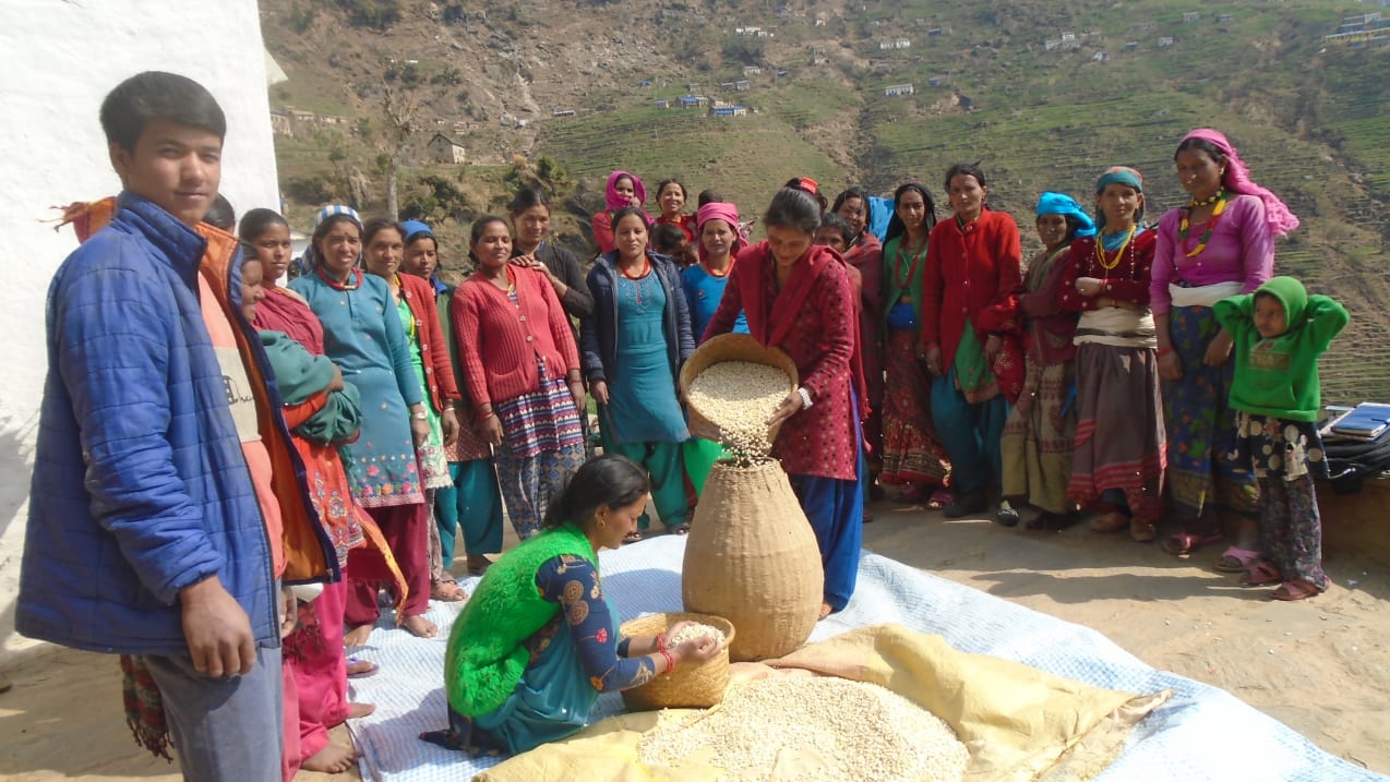 Two Nepalese women dressed in bright traditional clothing fill large storage containers with grain as a group of other women from their community watch. A large, steep mountain with some houses is behind them.