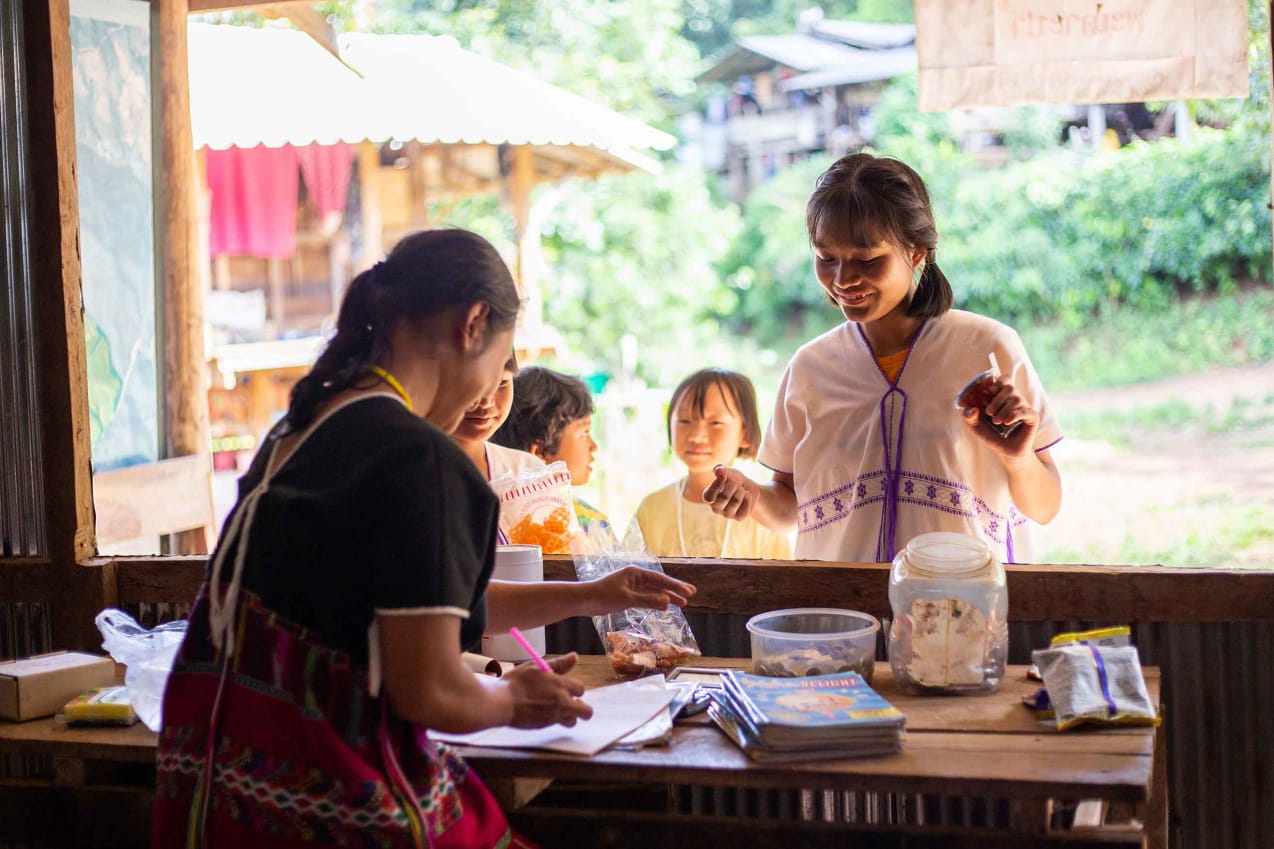 A Thai woman behind the counter of a shop writes down the detail of a sale as she serves a smiling female customer.