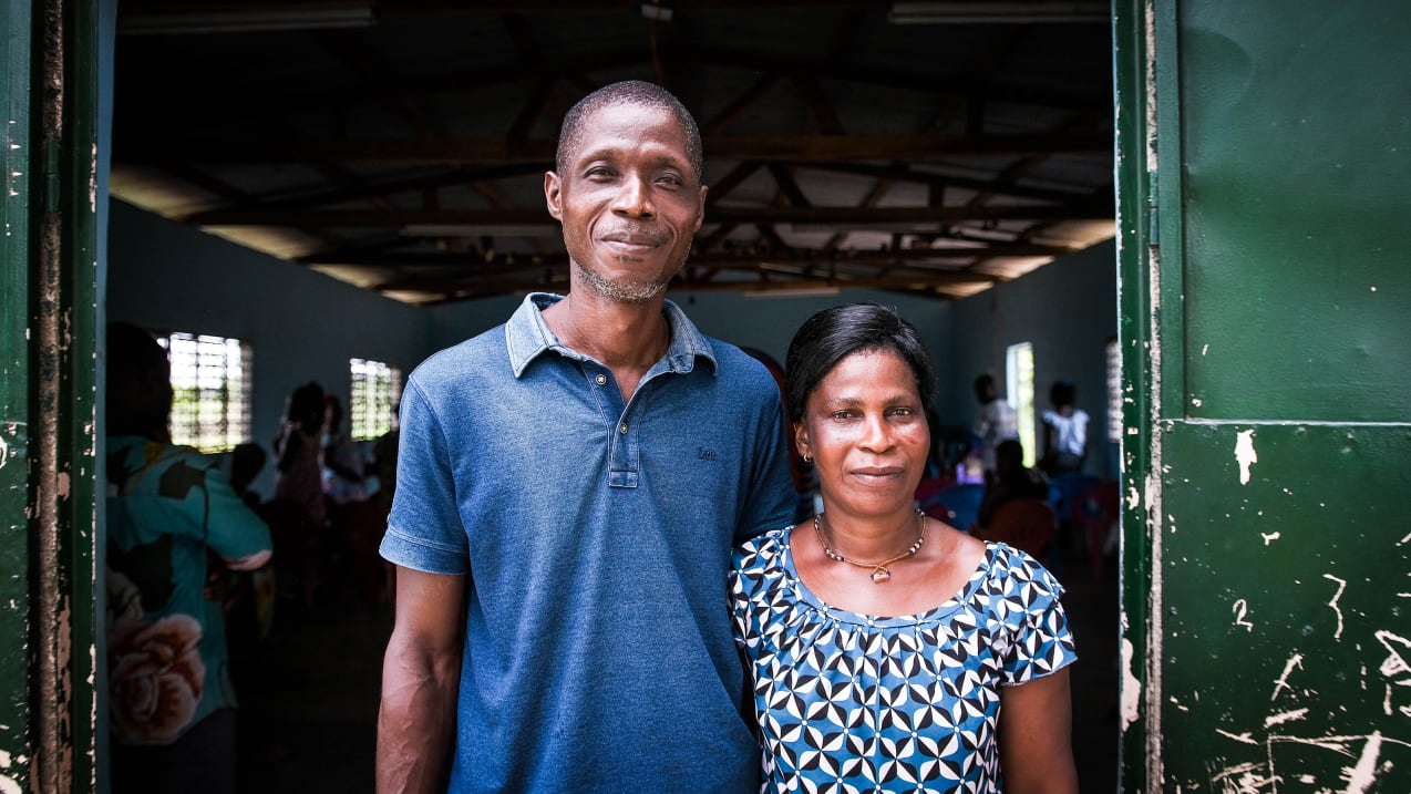 A man and woman stand in front of a church building open door with their arms around each other's backs.