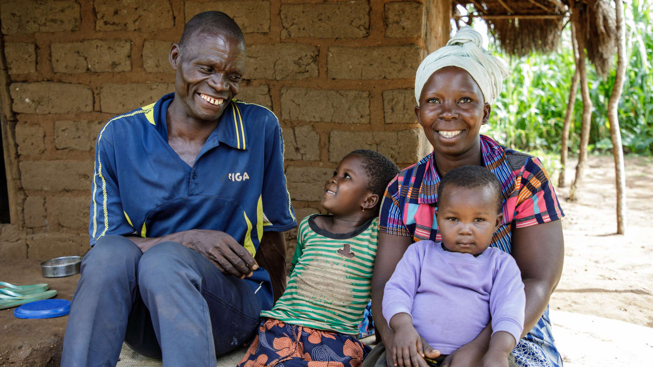 A man and woman are sat on the ground outside a buiding with their two children. The man smiles at the child sat next to him, while the child smiles back at him. The woman smiles at the camera, with a small child sat on her lap.