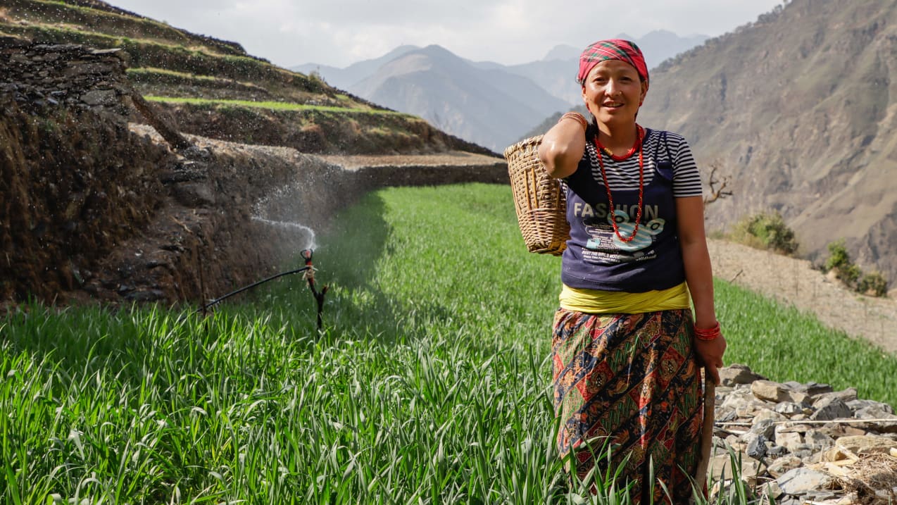 A woman is stood in a green field on a mountainside and smiles at the camera. 