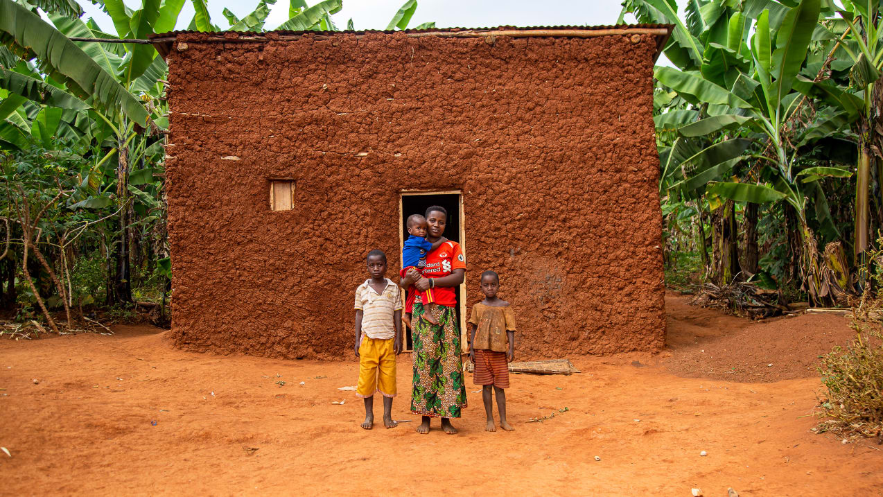 A woman stands in front of a house holding a young child in her arms, whilst two other children stand either side of her.