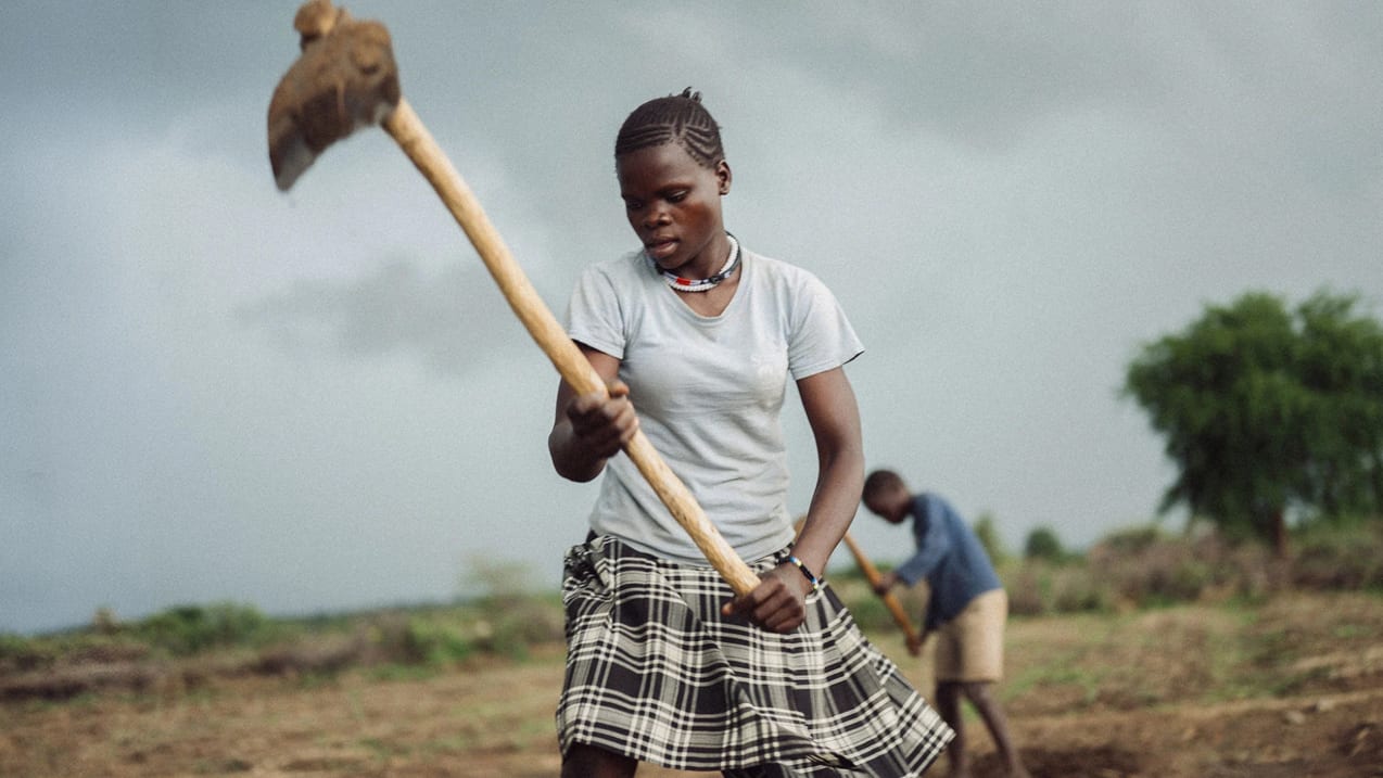 A young woman in a grey t-shirt and plaid skirt works a field with a hoe. In the blurry background, another person also uses a hoe under an overcast sky.