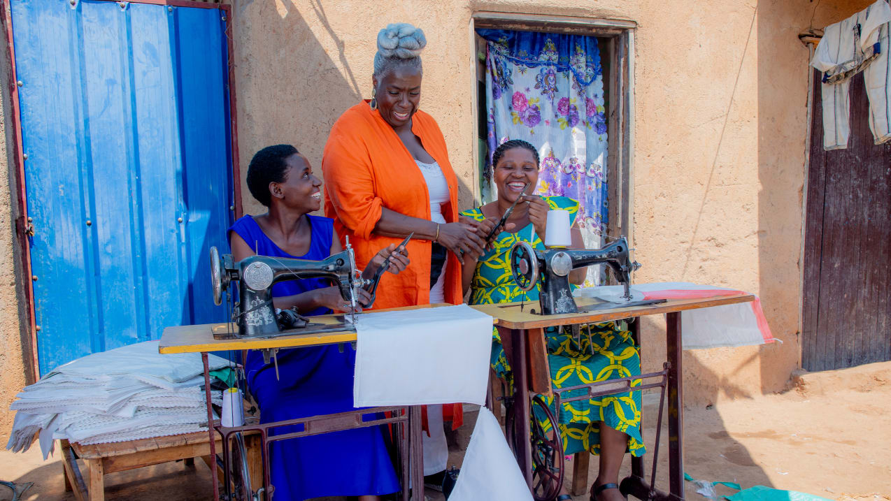 A woman stands in between two women sat at a table with sewing machines.