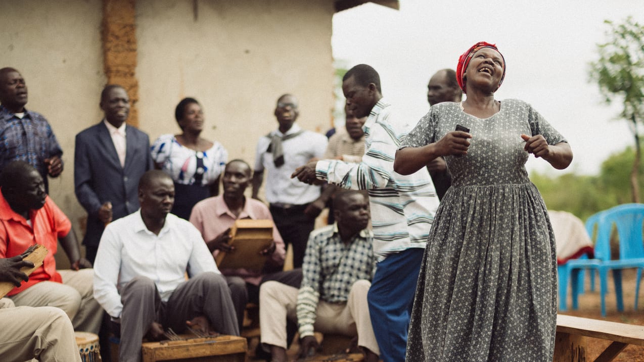 A group of people, mostly men, are seated outdoors playing musical instruments, while a woman in a patterned dress and red headscarf stands singing passionately.