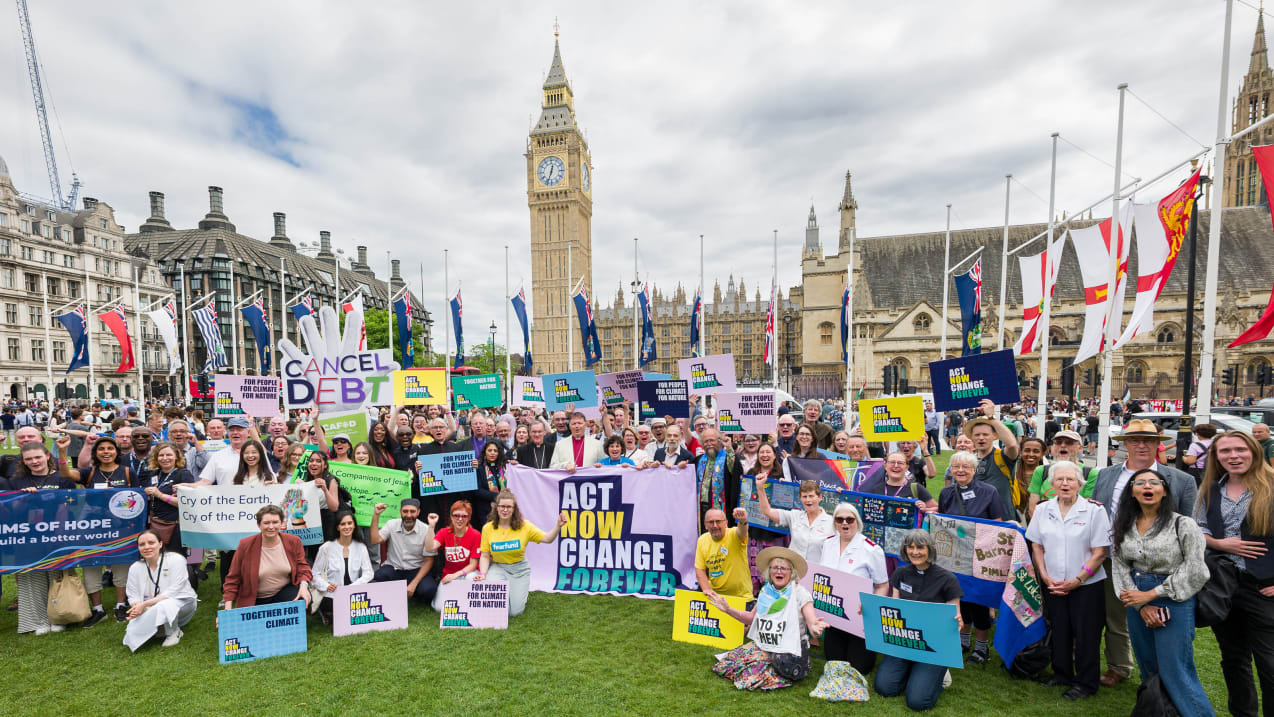 A large group of people holding signs and flags in front of Big Ben in London. One large sign says 'Act now change forever'.
