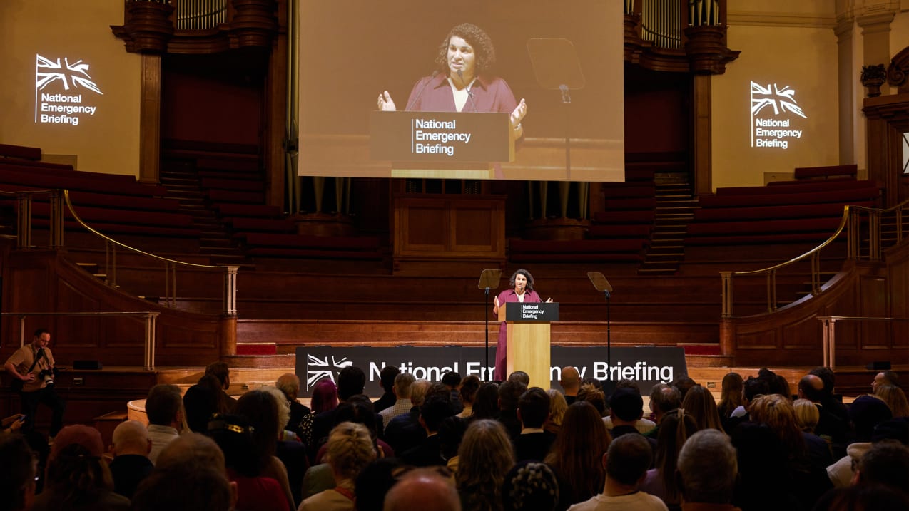 Woman speaks on a stage at the "National Emergency Briefing" to a crowd in auditorium, with a screen above.
