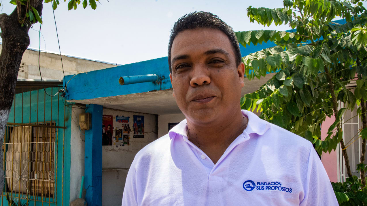 A man in a light polo shirt with a "Fundación Sus Propósitos" logo stands outdoors in front of a blue building and green trees.
