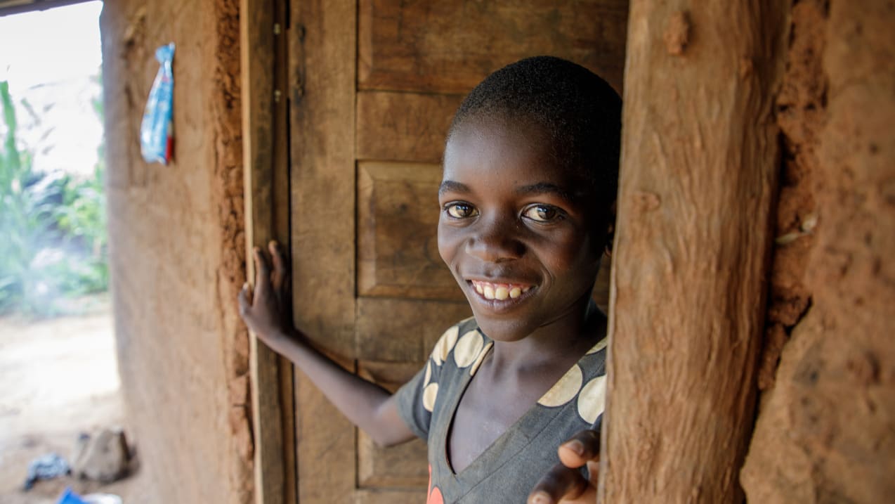 A young Black child with short hair smiles, looking towards the viewer, from a doorway in an earthen building. They wear a dark top with light polka dots.