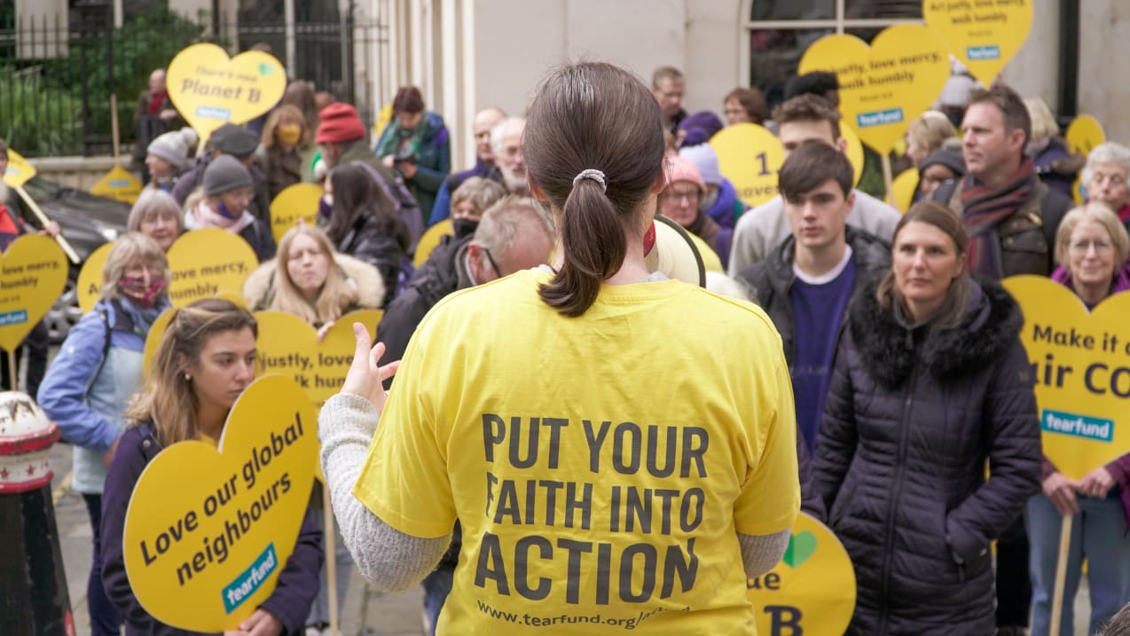 A crowd of people on a street, the camera is focused on the back of someone addressing the crowd, and on their t-shirt it says 'put your faith into action'.