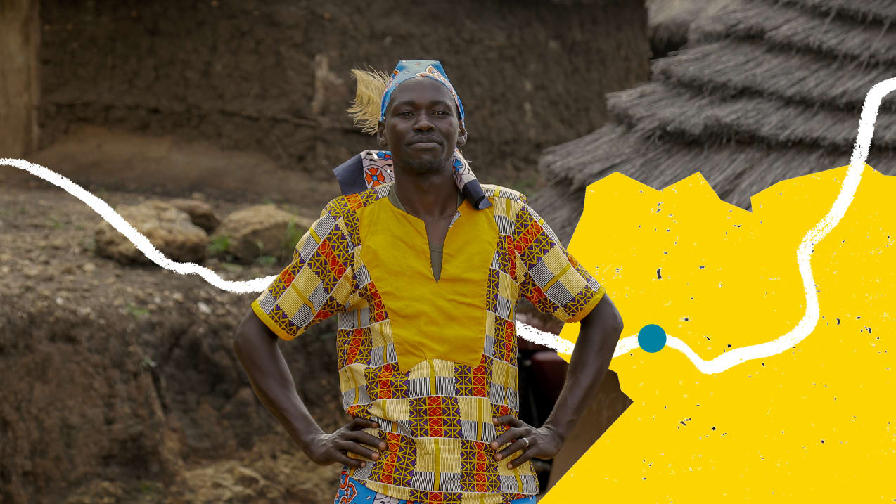 A man holds his hands on his hips and smiles at the camera. He is wearing a brightly patterned, yellow top and a scarf around his head with a large feather.