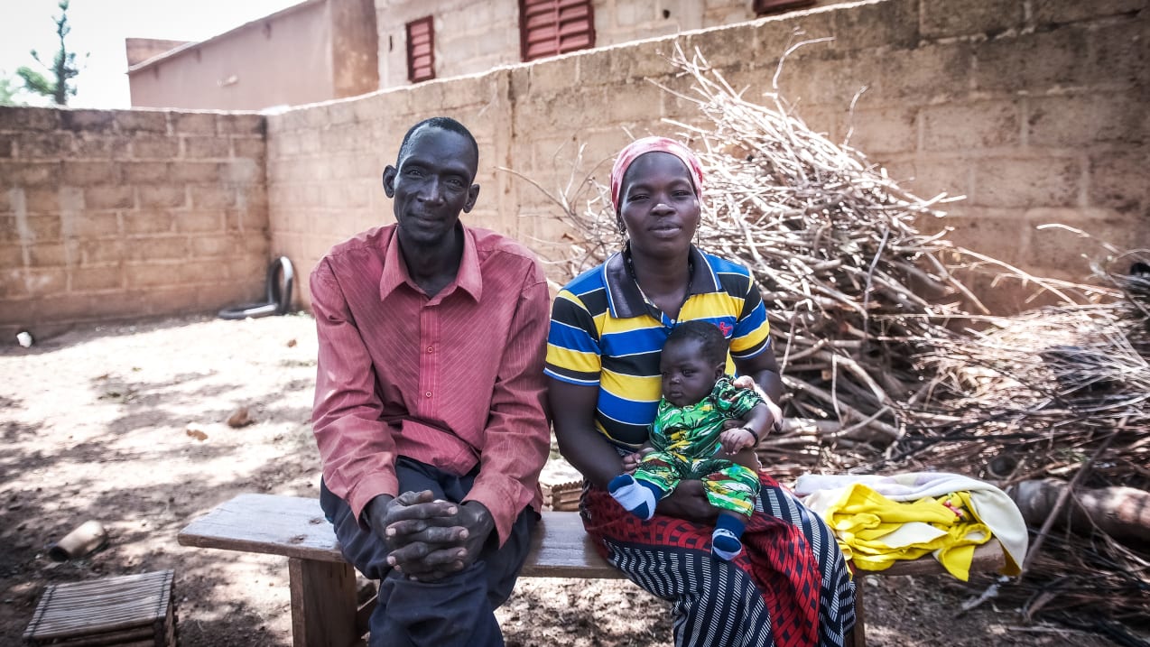 A man and woman sit on a bench smiling. The woman holds a baby on her lap.