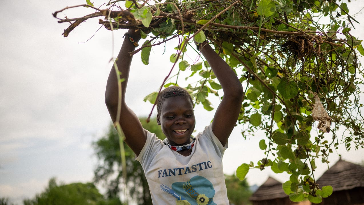 Esther, a young woman carries a large bunch of tree branches above her head and smiles. 