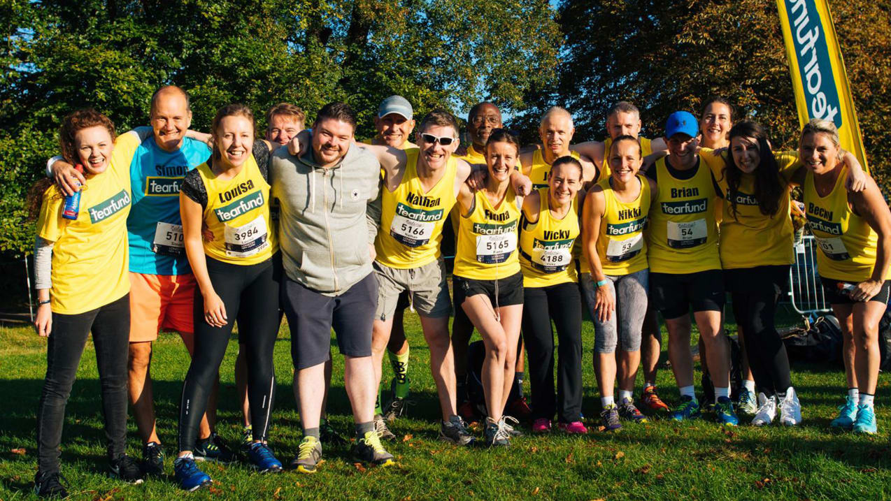 A large group of people all wearing yellow Tearfund t-shirts and running vests pose for a photo with their arms around each other.