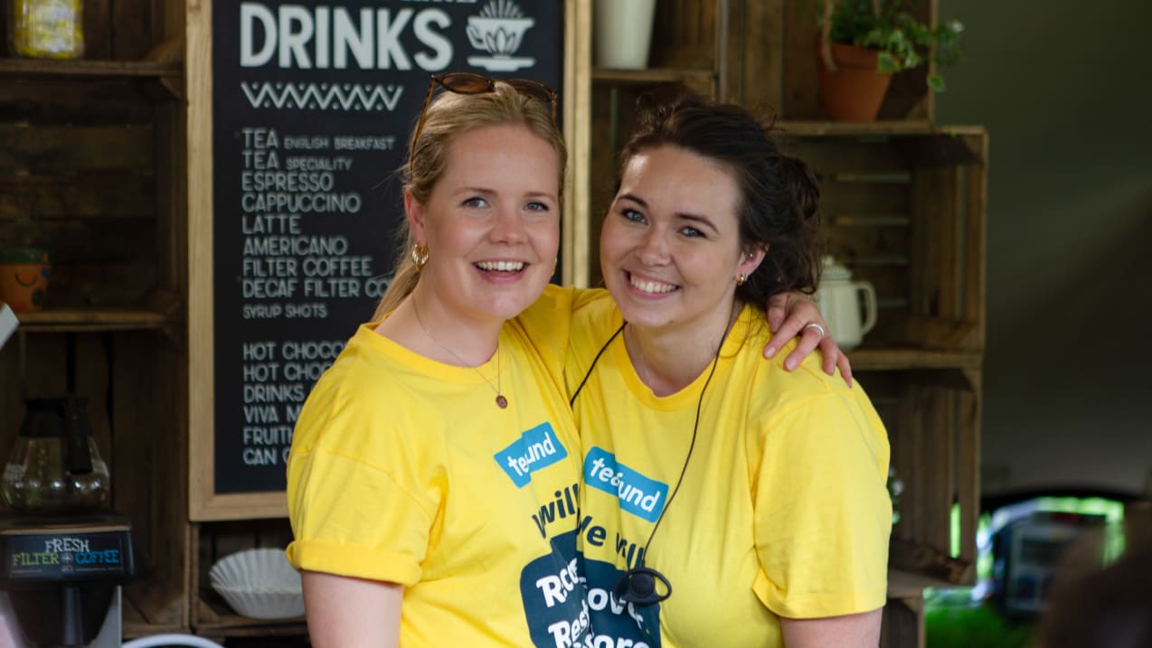 Two girls dressed in bright yellow Tearfund tshirts put their arms over each others shoulders and smile broadly to the camera. Behind them is a blackboard with the work 'Drinks' and a list of hot drinks.