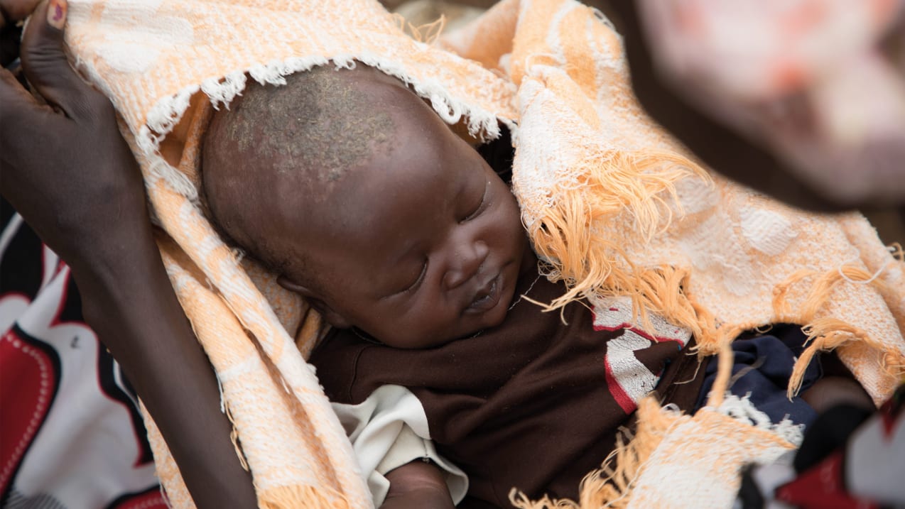 A baby with their eyes closed being held, dressed in a brown tshirt and wrapped in a woven cloth.