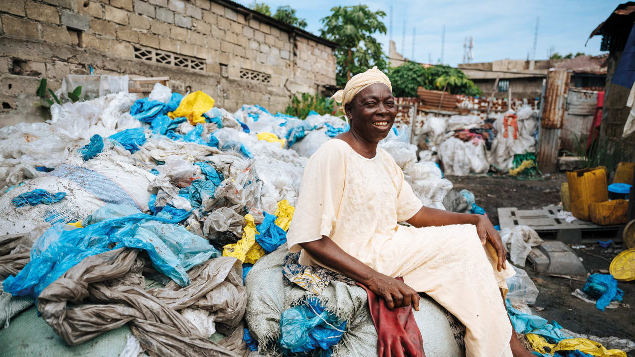A woman is sitting on top of a large pile of empty plastic bags, which line an alleyway wall, and smiles at the camera