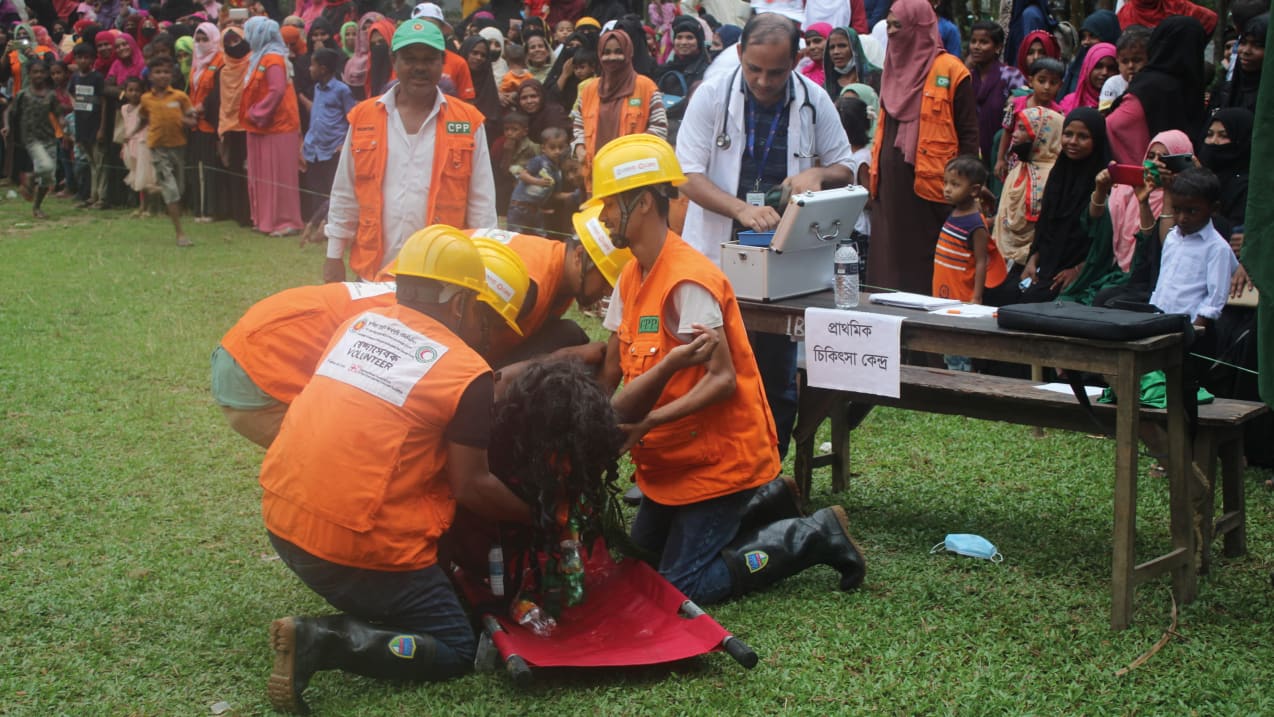 Four men in bright orange jackets help someone onto a red stretcher, while a crowd of people look on.