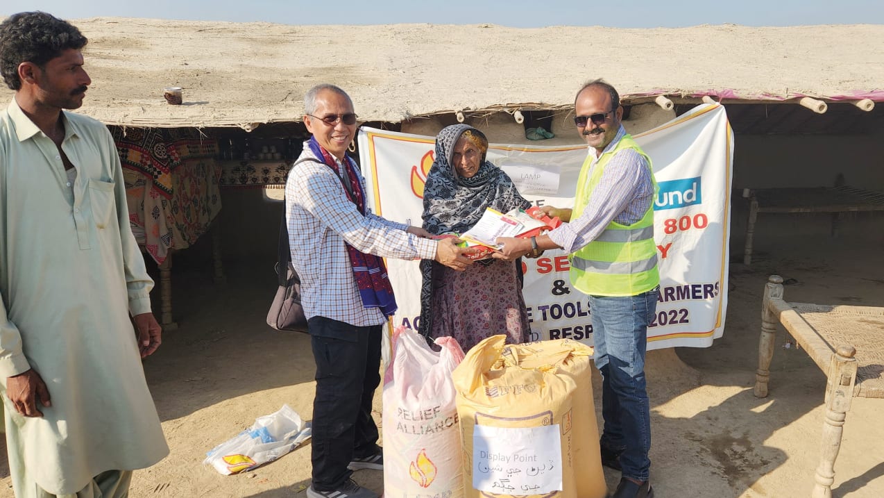 Two men either side of a woman, hold up a bag and a piece of paper. In front of them are two large sacks from the partner's relief work.