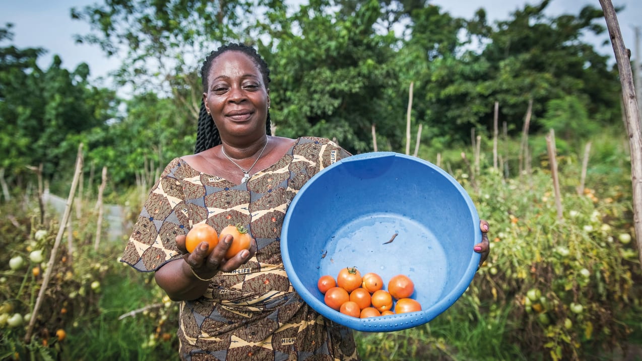 A woman holds a bright blue bucket with tomatoes inside, she holds two tomatoes in her other hand and smiles at the camera.