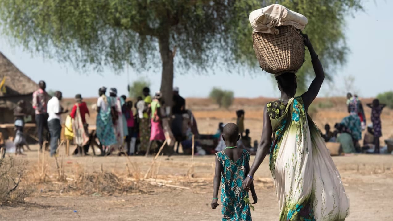A woman carrying a basket on her head, walking with a child toward a group of people congregating under a tree