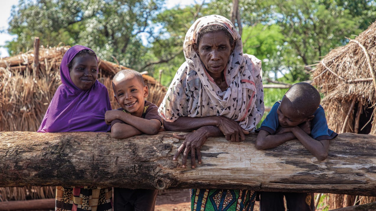 A woman and three children lean on a tree trunk in front of them, the children are smiling. Behind them are straw huts and trees.