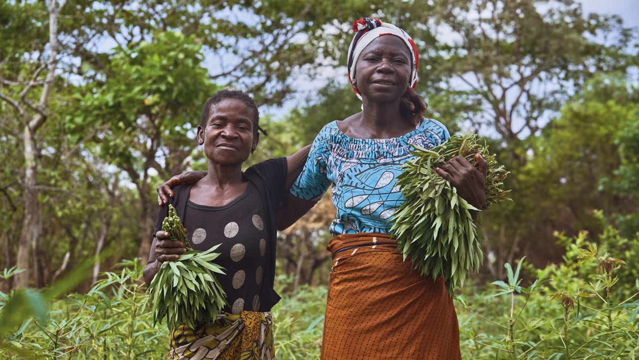 Two women put their arms around each other and hold crops in their other hand. Behind them are lots of trees and green shrubs.