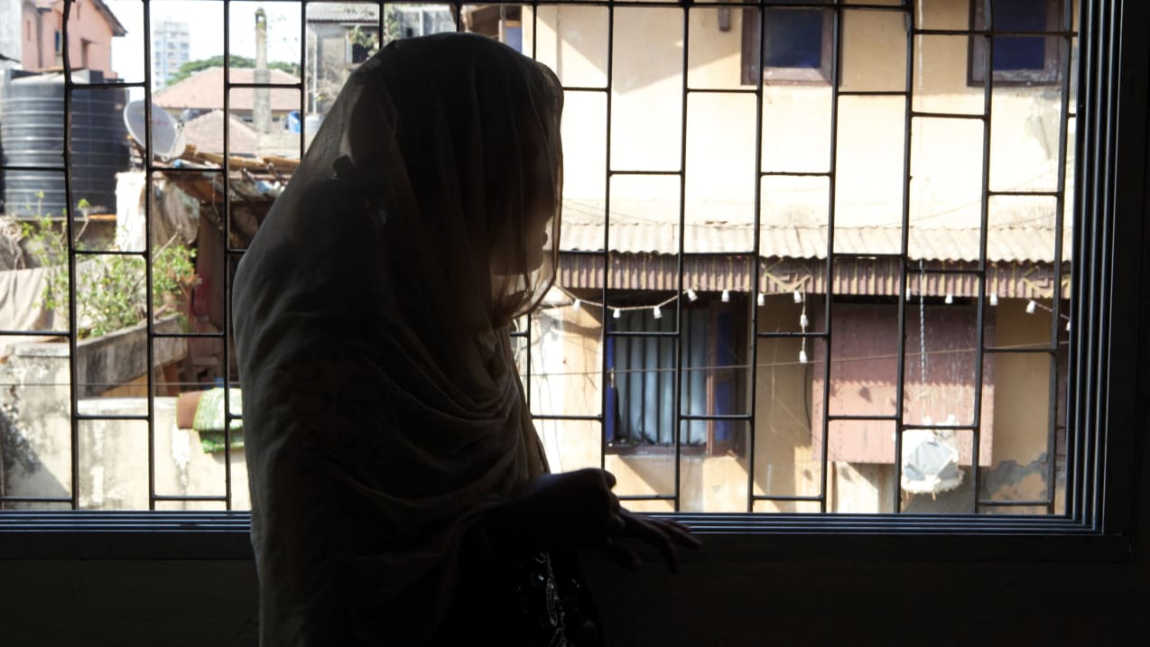 A shadow of woman stood in a dark room faces toward a bright window. She is wearing a scarf over her head and body.