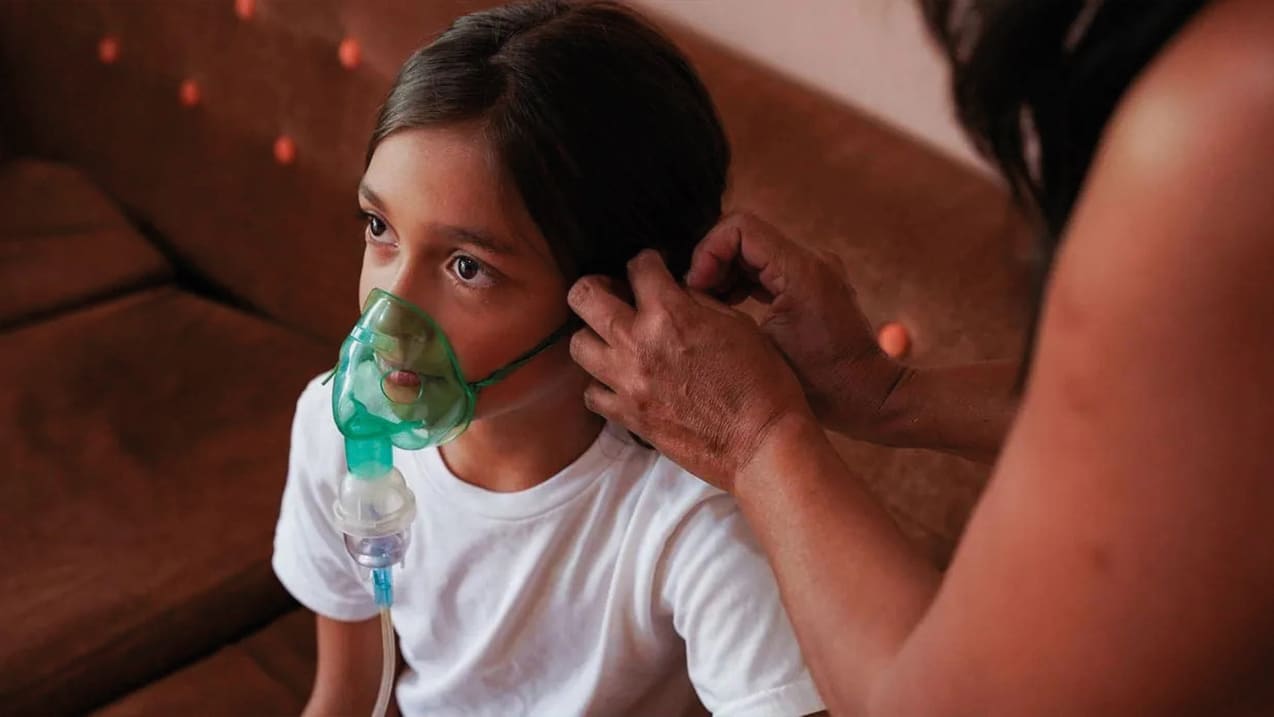 A young girl is sat down and looking up, as someone puts a nebuliser mask on her.