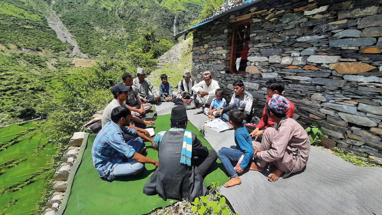 A group of people are sat on the floor outside talking They are sat next to a stone building and to the other side of them you can see steep green hillsides.