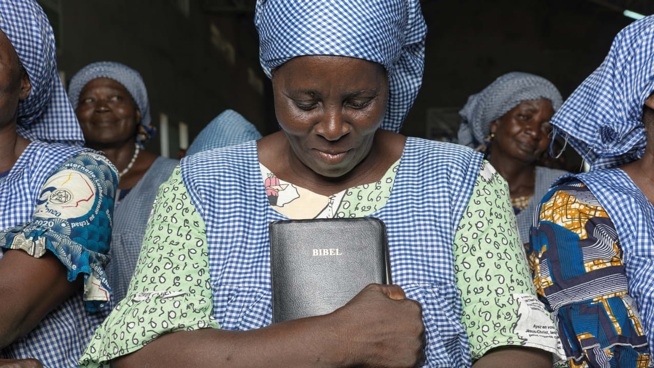 A woman looks down and clutches her bible to her chest. Around her are several other women.