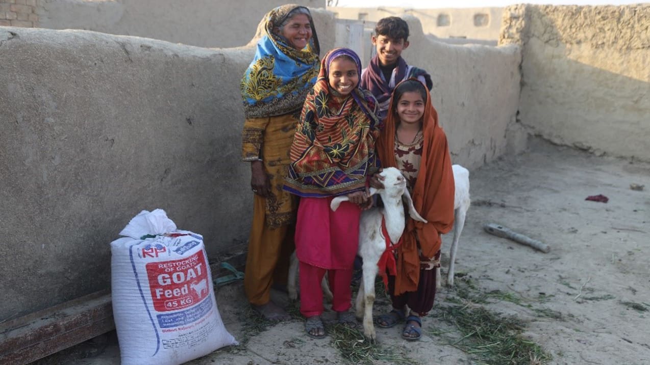 A woman stands with her three children and their goat. Beside them is a sack of goat feed.
