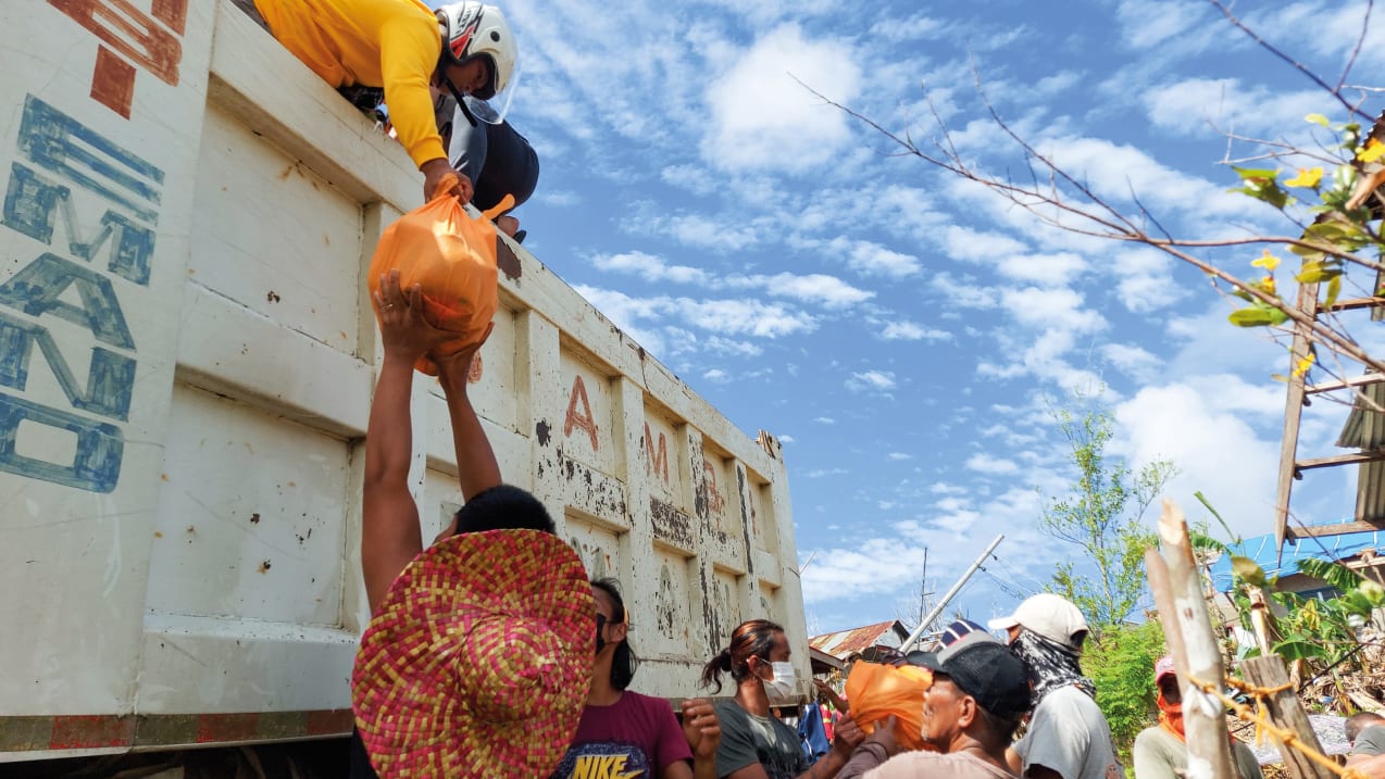 Someone stood in the back of a large truck passes an orange plastic bag down to someone stood on the ground.  Several other people are stood on the ground passing plastic bags.