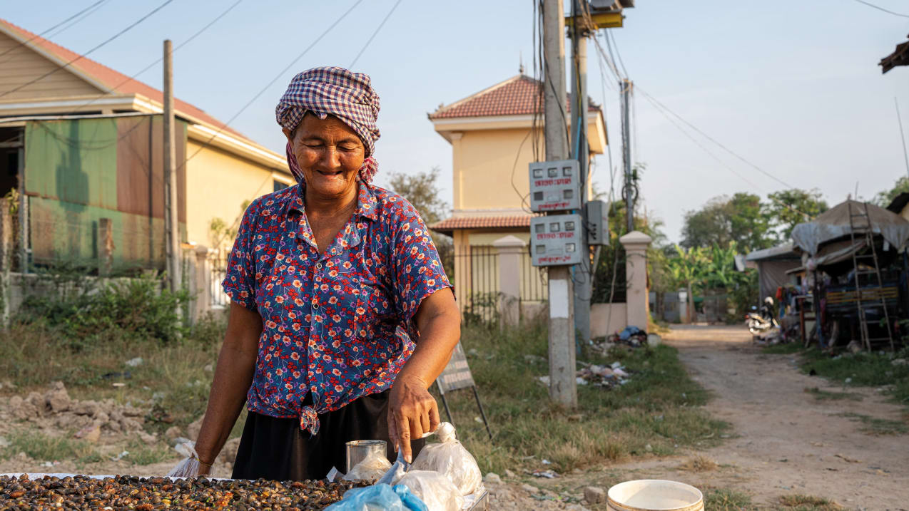 A smiling woman in a floral shirt and checkered headscarf stands at an outdoor food stall in a residential area with power lines and buildings in the background.