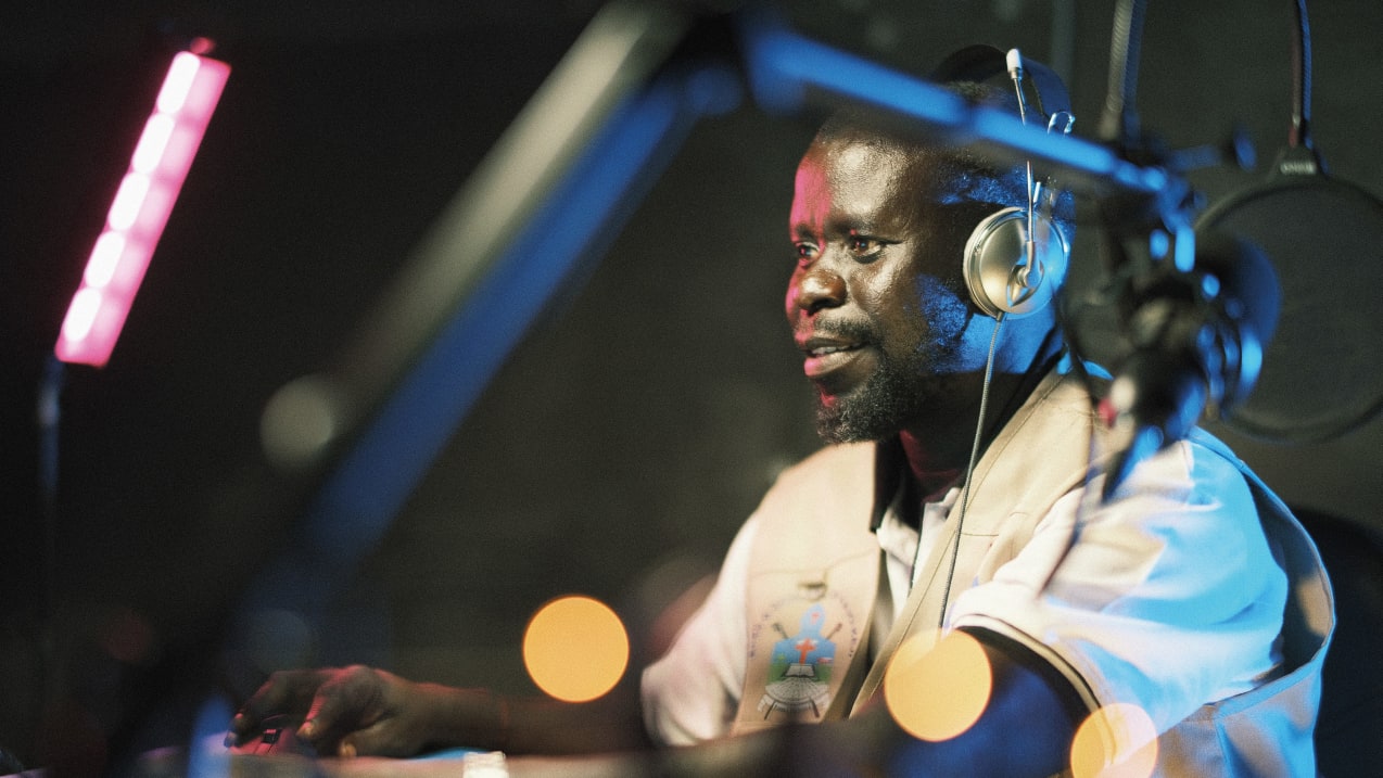 A man wearing headphones sits in a dimly lit recording studio, speaking into a microphone with colorful lights and audio equipment around him.