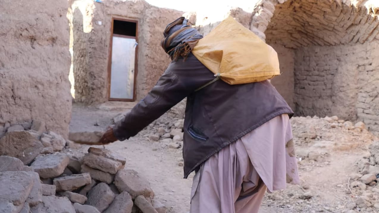 Someone dressed in a coat and headscarf, with a yellow bag strung to their back moves a brick to the top of a pile of them. In the background is a building and rubble.