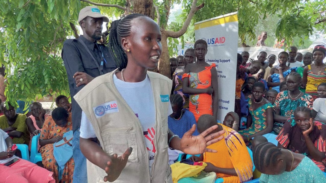 A woman stands in front of a large group of people sat down. She is wearing a vest jacket with Tearfund and USAID logos on and gestures with her hands. Behind her is a man wearing a cap with the Tearfund logo on.