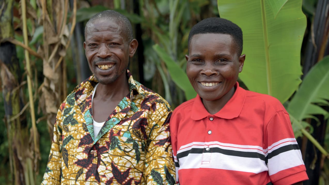 A couple stand next to each other smiling. Behind them are lots of green vegetation.