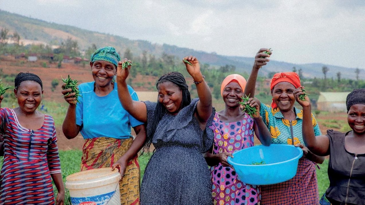 A group of women hold up green beans in their hands and smile. Two of them hold large plastic containers filled with beans.